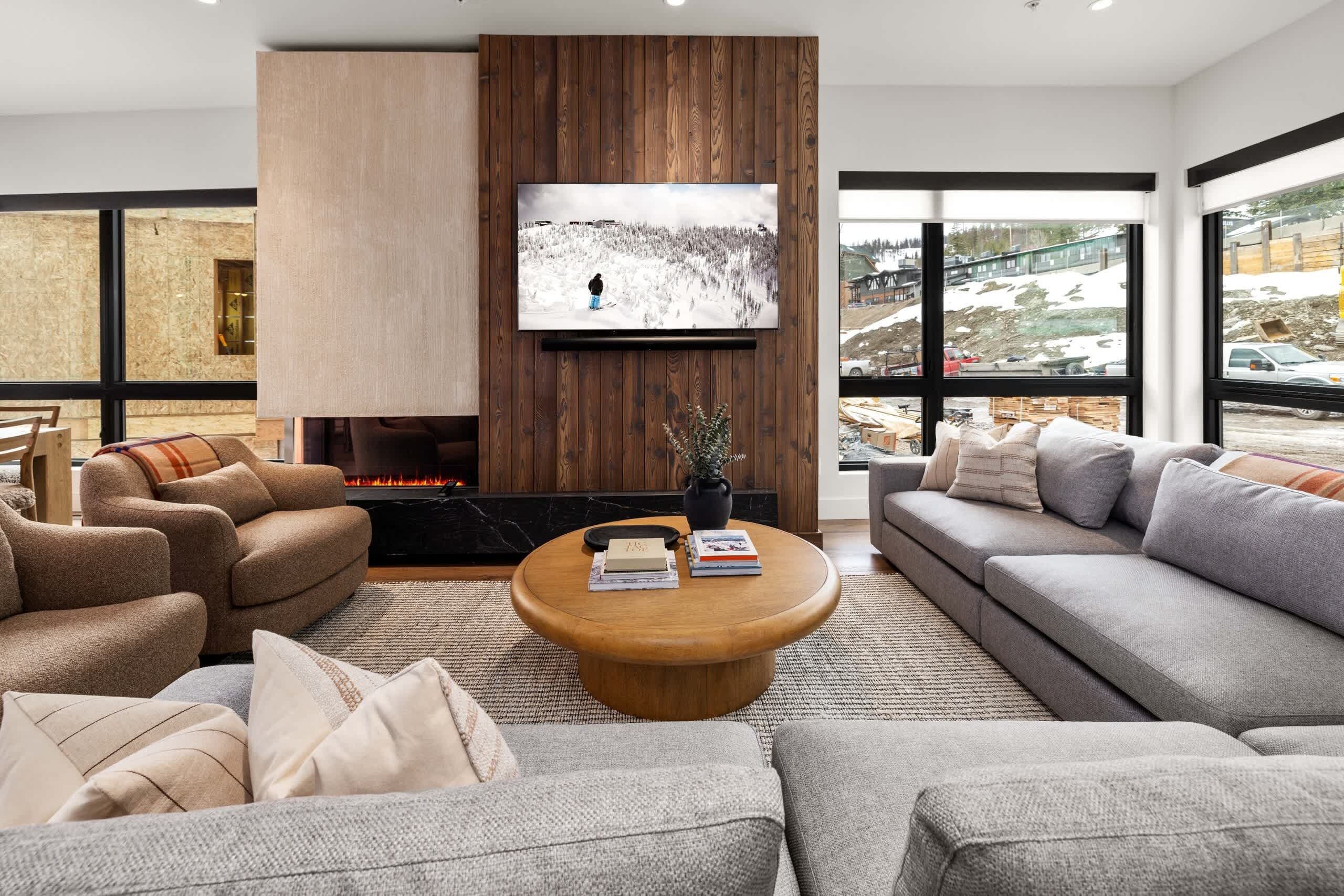 A modern living room with a fireplace, a large TV mounted on a wooden wall, and a view of a snowy landscape through large windows.