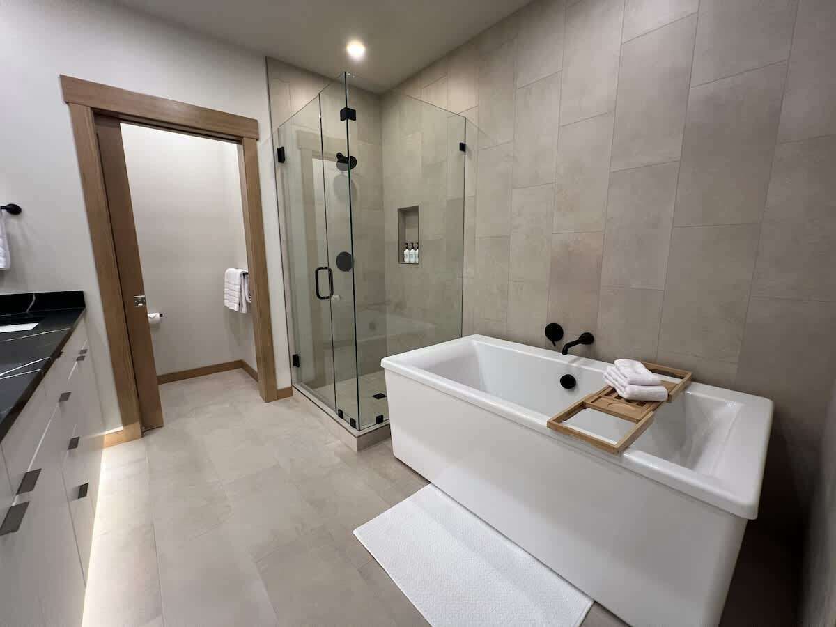 A modern bathroom with a glass-enclosed shower, a white bathtub, and a wooden tray.