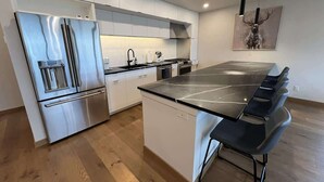 A modern kitchen with a stainless steel refrigerator, a central island with bar stools, white cabinetry, and a painting of a deer on the wall. - Clubhouse & Pool Amenities, 40 Mins to Glacier NP (Whitefish)