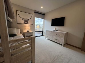 Ein Schlafzimmer mit Bett, Nachttisch, Lampe, einem an der Wand montierten Fernseher, einer Kommode und einem großen Fenster mit Blick auf die Stadt.