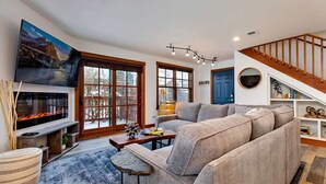 A modern living room with a large flat-screen TV, a sectional sofa, a fireplace, and a staircase. - Downtown Condo, Sleeps 7, 35 Mins to Glacier Park (Whitefish)