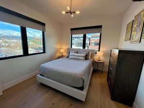 Un dormitorio con una cama grande, dos mesitas de noche, un tocador y vistas a un paisaje urbano a través de amplias ventanas.