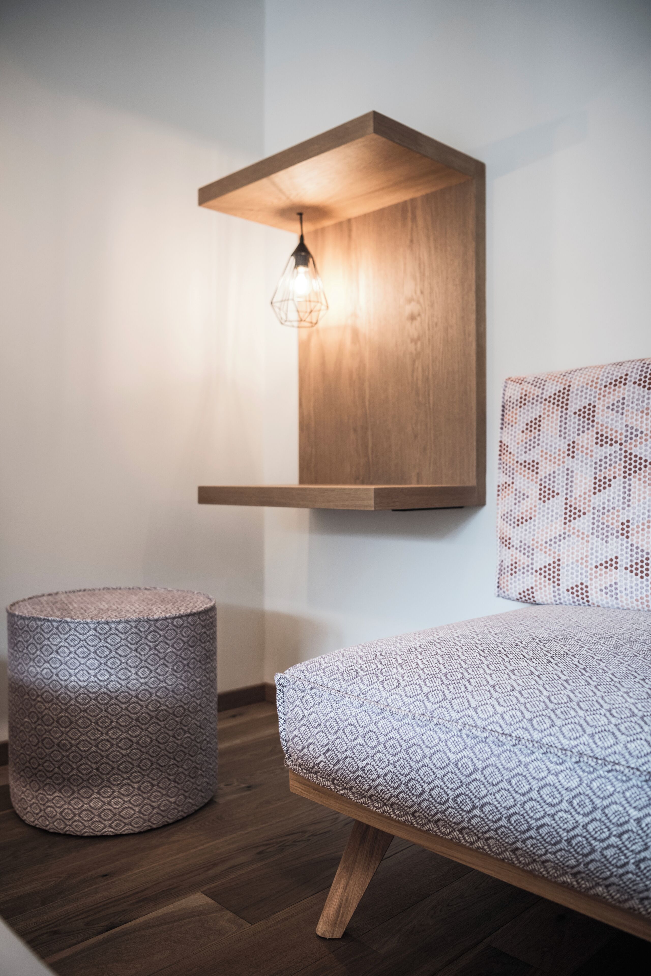 A modern bedroom with a wooden headboard, a patterned bedspread, a round ottoman, and a wall-mounted light fixture.