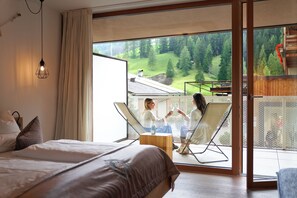 Un dormitorio con una cama grande, un espacio de descanso con dos sillones y vista a un paisaje boscoso a través de un ventanal amplio.