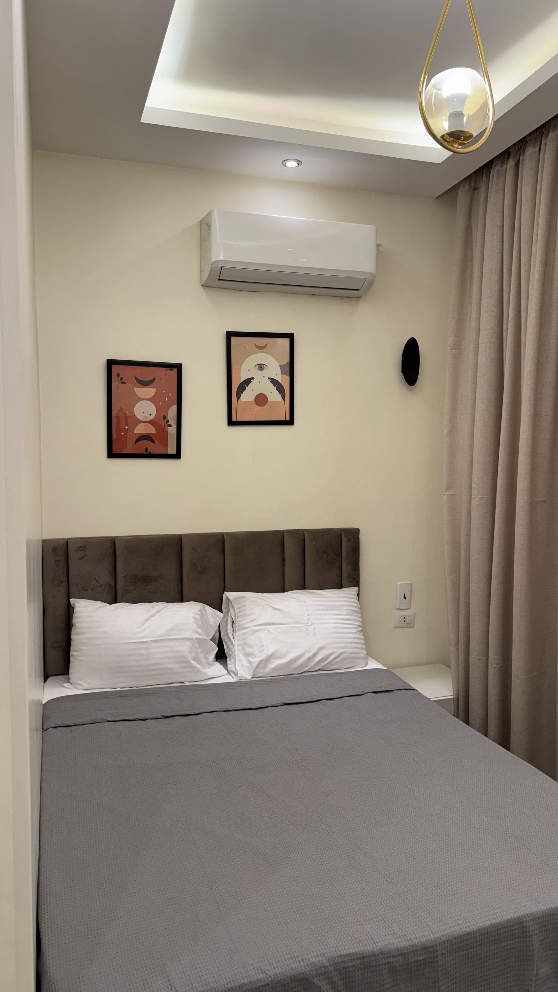 A bedroom with a bed, a ceiling light, an air conditioner, and two wall art pieces.