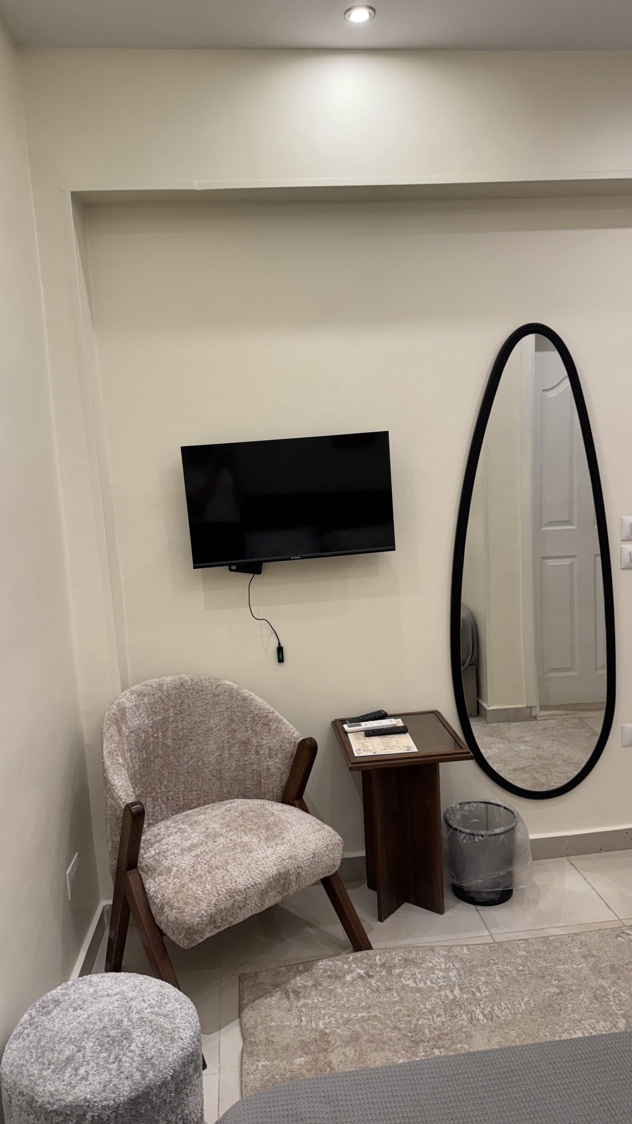 A modern living room with a flat-screen TV mounted on the wall, a large mirror, a cushioned armchair, a small wooden side table, and a trash can.