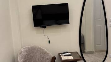 A modern living room with a flat-screen TV mounted on the wall, a large mirror, a cushioned armchair, a small wooden side table, and a trash can.