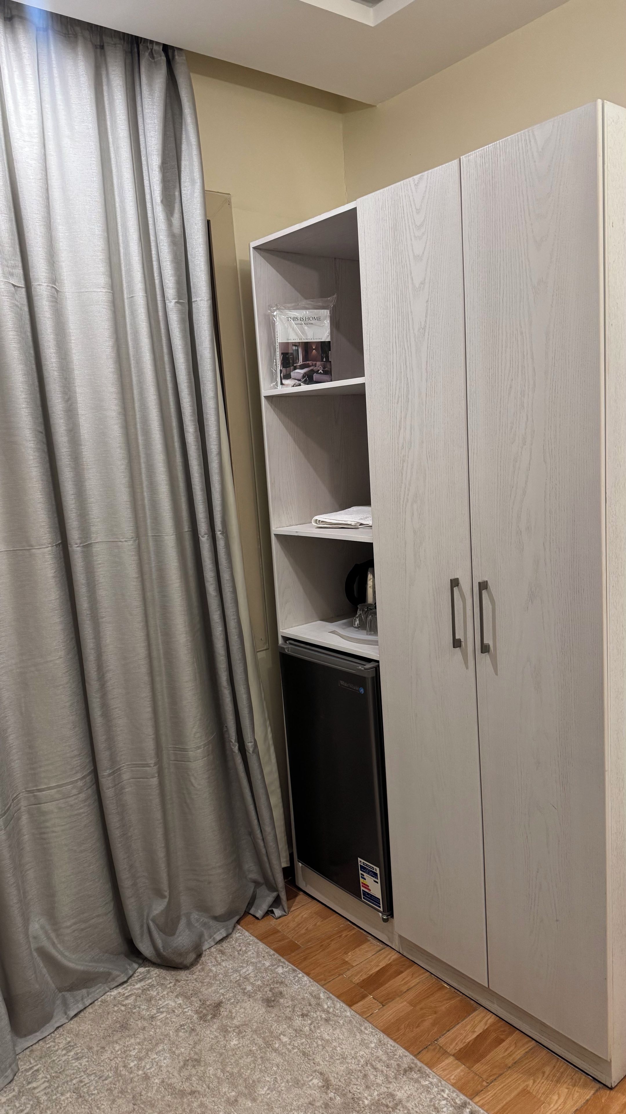 A room with a grey wardrobe, a small black appliance, and a grey curtain.