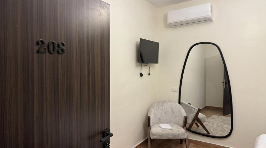 Ein Zimmer mit Holztür mit der Aufschrift '208', ein grauer Sessel, ein Flachbildfernseher an der Wand und ein großes Spiegel.