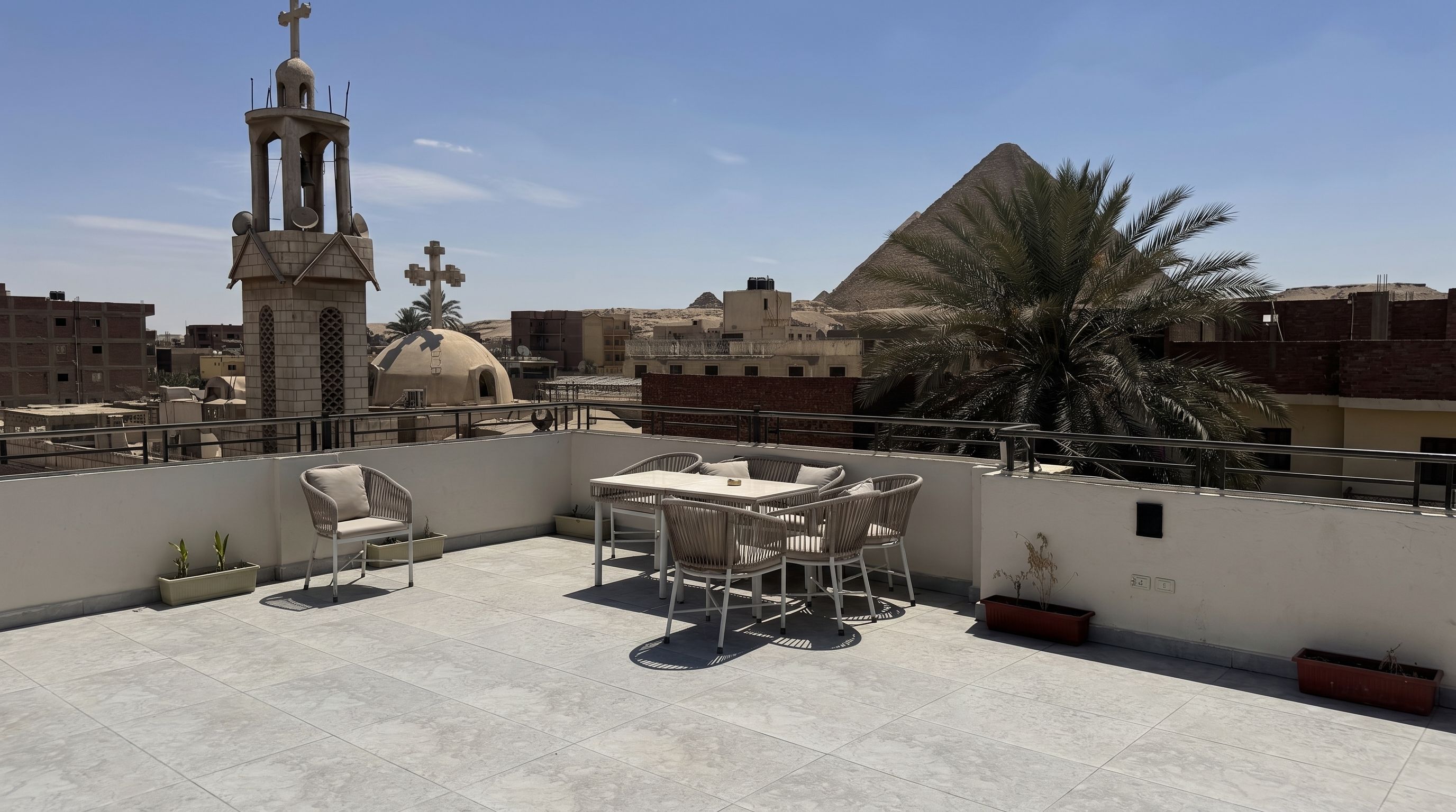 A rooftop terrace with a table and chairs, a bell tower, and a pyramid in the background.