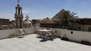 A rooftop terrace with a table and chairs, a bell tower, and a pyramid in the background.