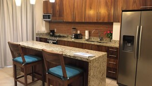Una cocina moderna con encimeras de granito, armarios de madera, un refrigerador de acero inoxidable y dos taburetes de bar con cojines azules.