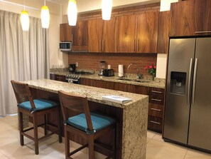 A modern kitchen with granite countertops, wooden cabinets, a stainless steel refrigerator, and two bar stools with blue cushions. - Cozy Condo with Stunning Infinity Pool (Puerto Vallarta)