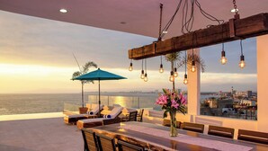 Terraza en la azotea con vistas panorámicas de la ciudad, una mesa de comedor de madera, sillas de lounge y lámparas colgantes.