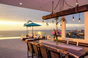 A rooftop terrace with a panoramic city view, a wooden dining table, lounge chairs, and hanging light fixtures. - Cozy Condo with Stunning Infinity Pool (Puerto Vallarta)