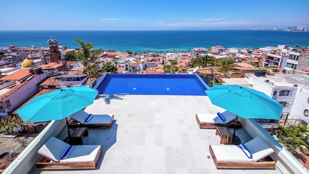 Una piscina en la azotea con dos sombrillas azules, sillas de descanso y vistas al mar y a la ciudad de abajo.