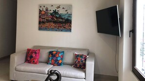 Un salón con un sofá gris, cojines coloridos, un televisor montado en la pared y un cuadro de una playa.