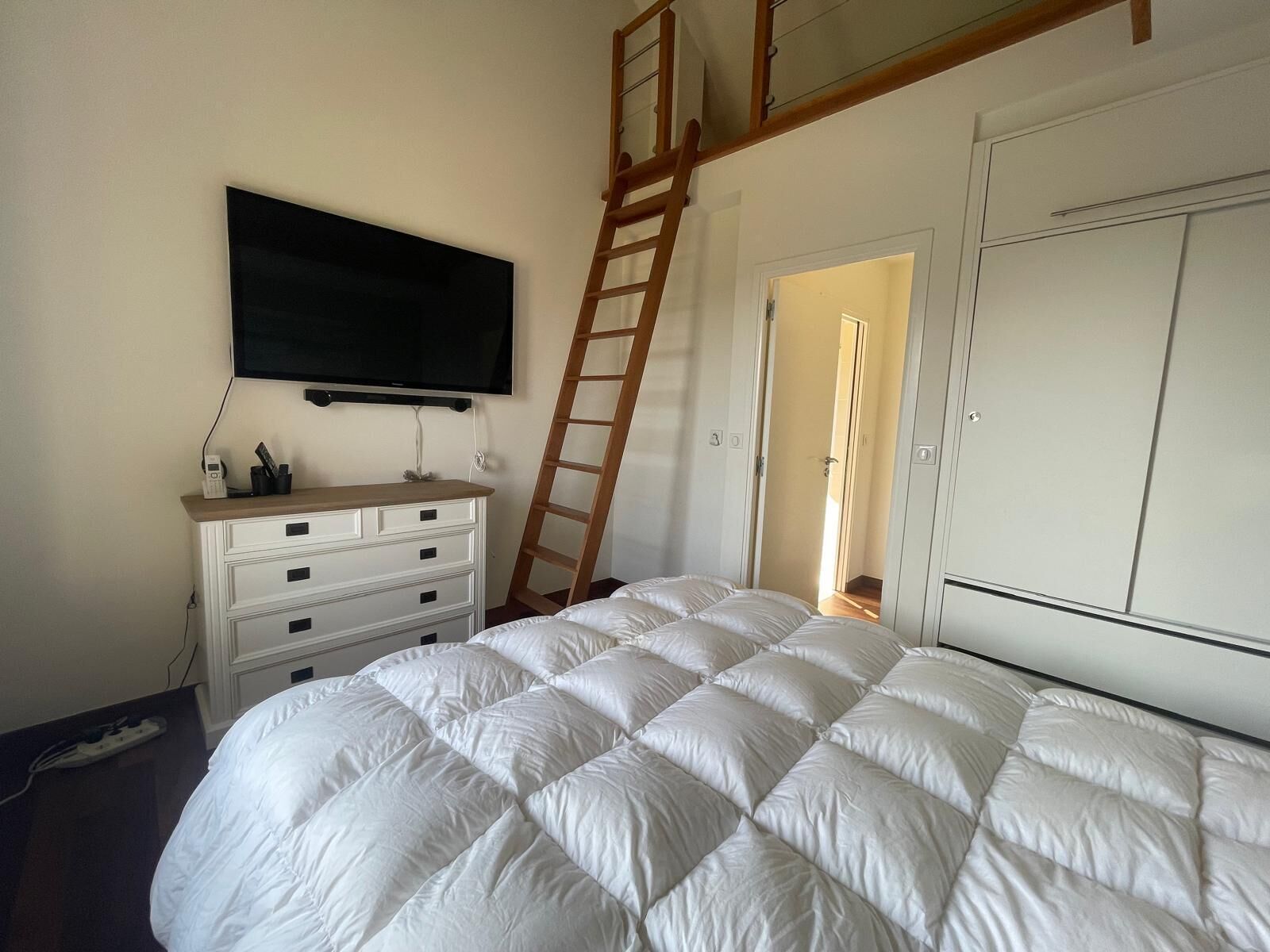 A bedroom with a bed, a TV mounted on the wall, a dresser, a ladder, and a wardrobe.