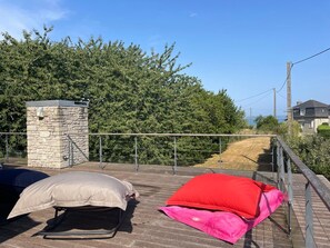 A wooden deck with two pillows, a stone wall, and a metal railing overlooking a landscaped area. - VILLA BERGEONS NEAR THE BEACH CENTER IN ST CAST—4-STAR TOURISM RATING (Saint-Cast-le-Guildo)