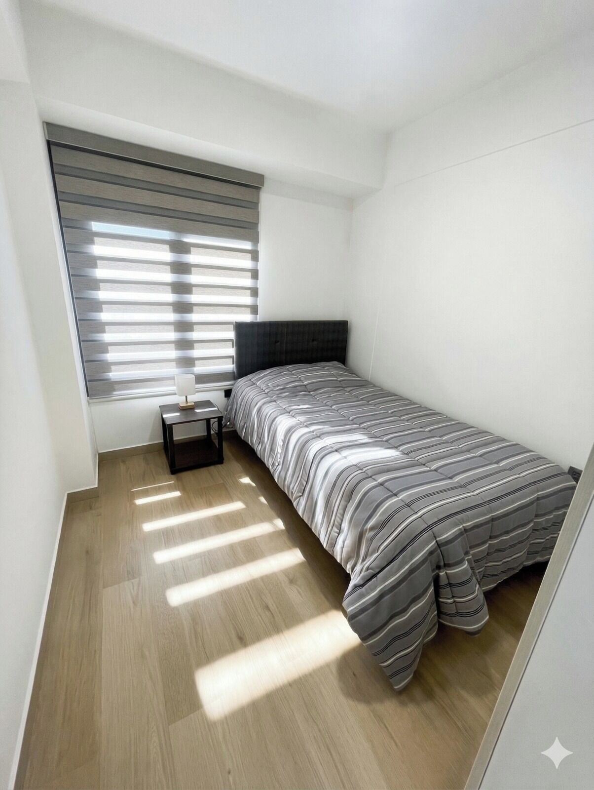A small, single-bed room with a sloped ceiling, a window with blinds, a bedside table, and a light-colored wooden floor.