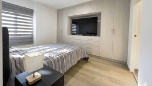 A bedroom with a bed, a TV mounted on the wall, a desk with a lamp, and a window with blinds. - Apartment in Yanahuara: Modern, Central, and Fully Equipped (Yanahuara)