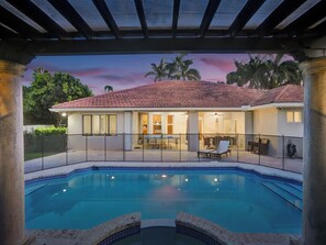 Una piscina con bordeado de baldosas, un porche cubierto con mobiliario exterior, y una casa con tejado de baldosas y grandes ventanales.