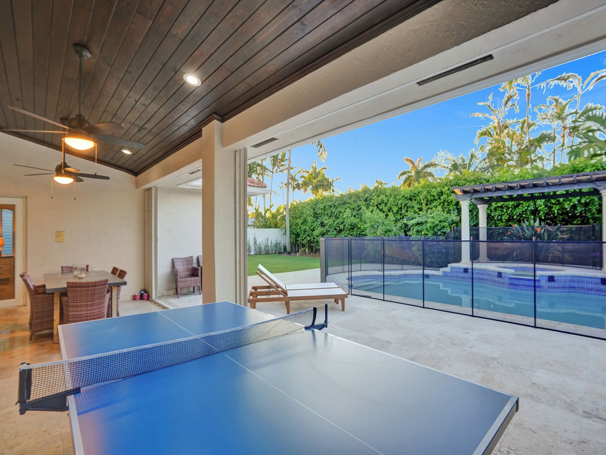 An indoor ping pong table with a glass enclosure, a patio area with chairs, and a view of a swimming pool and greenery.