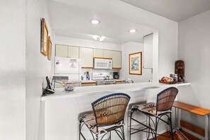 A compact kitchen with white cabinets, a microwave, and a coffee maker. There are two wicker chairs and a wooden table. - Kihei Bay Vista C-201 - Kihei 1BR Condo, AC, Lanai, Wifi, 45 Sec to Sugar Beach! (Kihei)