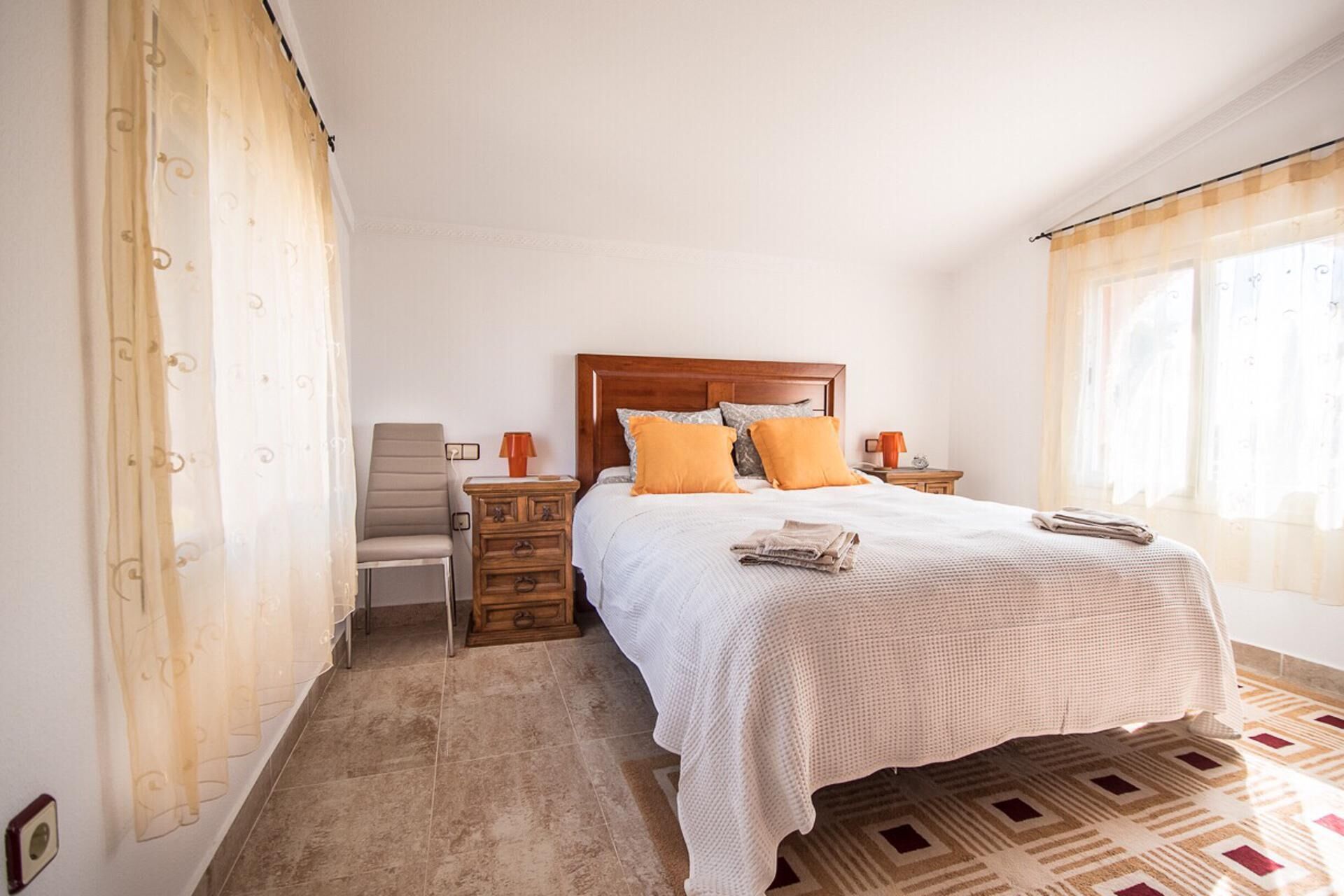 Une chambre à coucher avec un lit, une chaise, une table de chevet et une fenêtre avec des rideaux.