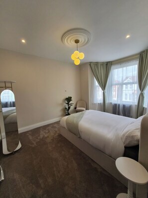 A bedroom with a bed, a bedside table, a mirror, a plant, and a window with curtains. - Apartment Sleeps 4 With Parking (Ilford)