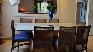 Un coin repas avec une table blanche, des chaises bleues, un vase bleu contenant des plantes, un ventilateur de plafond et une fenêtre avec des stores.