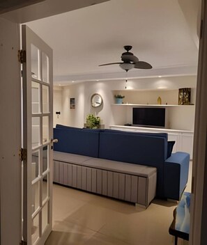 Ein Wohnzimmer mit einer Couch, einem Fernseher, einem Deckenventilator und einer Tür zu einem anderen Raum.
