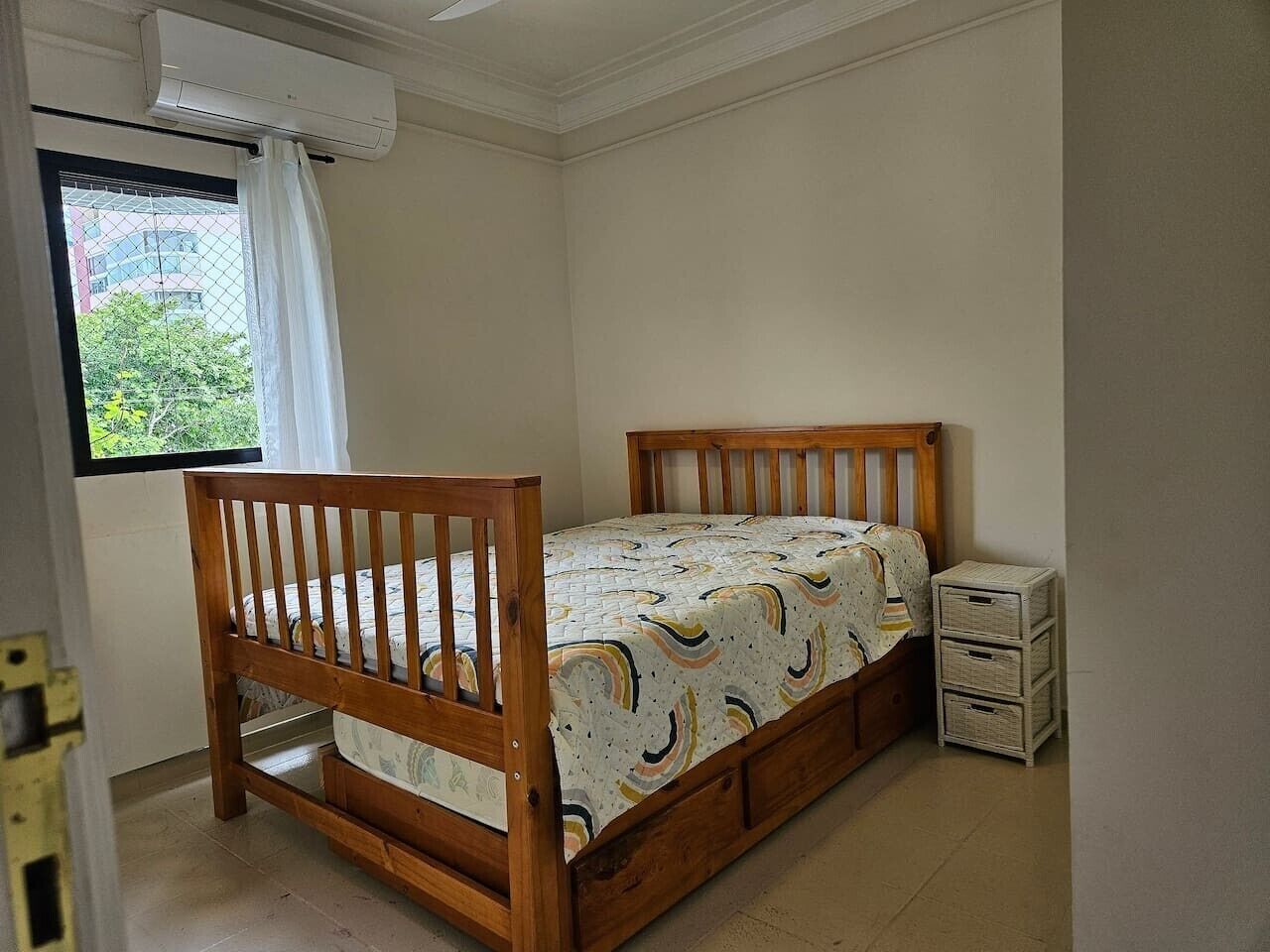 A bedroom with a wooden bed, a window with a curtain, and a small white cabinet.