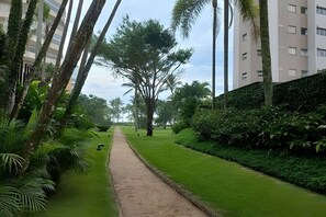 A pathway flanked by lush greenery and tall trees, leading towards a modern building with multiple floors. - Cozy beachfront apartment in Module 2 (Riviera de São Lourenço)