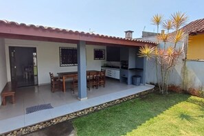 A covered patio with a dining table and chairs, a kitchen area, and a small garden with a palm tree. - Bwelcome - Casa Indaiá: Espaço Gourmet e Brisa Mar (Bertioga)