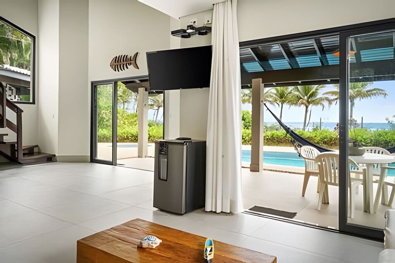 A modern living room with a flat-screen TV, a refrigerator, a wooden coffee table, and a view of a pool and palm trees.