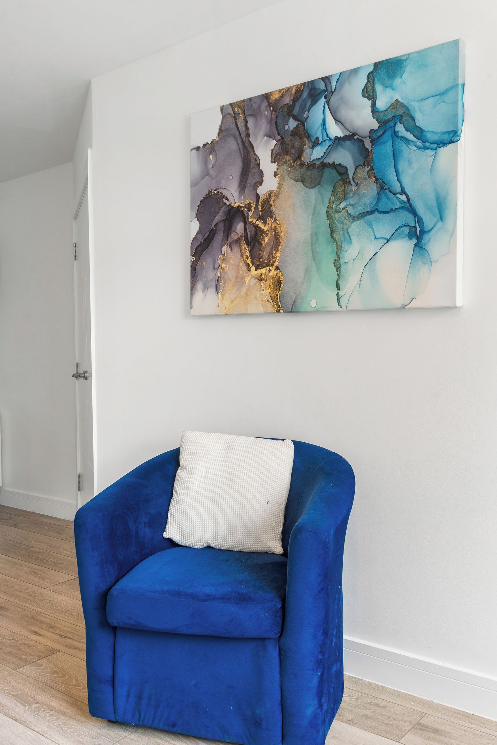 A room with a blue armchair, a white cushion, and a large abstract painting on the wall.