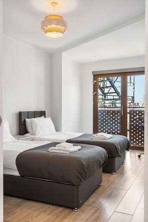 A modern bedroom with two beds, a large window, and a view of an industrial area. - Penthouse for 8, Roof Terrace, 9 Min Drive to Studio Tour (Hertfordshire)