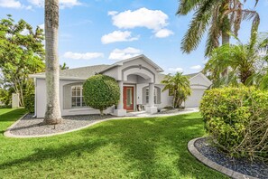 A single-story house with a red door, white exterior, and a well-maintained lawn. - Envision Paradise: Gorgeous 4 Bedroom Waterfront Home with Private Pool/Spa and Large Kitchen (Cape Coral)