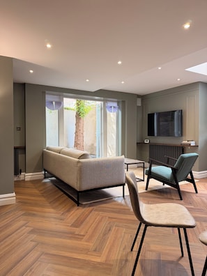 Uma sala de estar moderna com um sofá, duas poltronas, uma televisão e uma janela grande com vista para uma árvore.