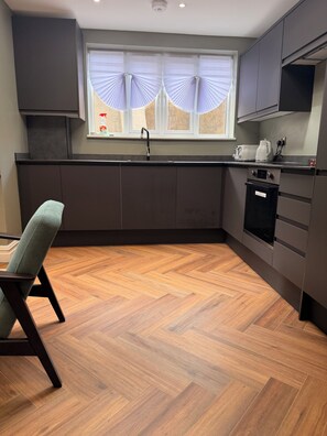 Uma cozinha moderna com armários escuros, piso de madeira em padrão herringbone, janela com cortinas translúcidas e forno embutido.
