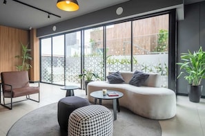 A modern living room with a sofa, ottoman, and chair, a round coffee table, and large windows with a view of the outside. - Luxury Stay Steps from Larcomar | Pool & Parking (Miraflores)