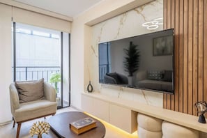 A modern living room with a sofa, a coffee table, a flat-screen TV mounted on a marble wall, and a balcony with a railing. - Luxury Stay Steps from Larcomar | Pool & Parking (Miraflores)