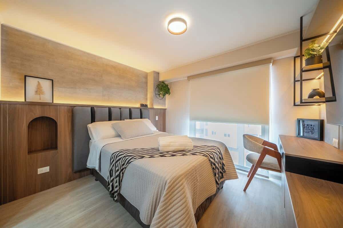 A modern hotel room with a bed, a desk, a chair, a plant, and a window.