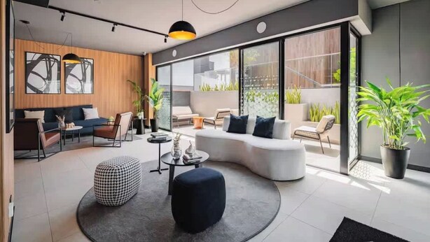 A modern living room with a curved sofa, a round coffee table, and a black ottoman. There are two chairs, a small table with decorative items, and a large window with a view of greenery outside. - Steps from Larcomar | 3BR · A/C · Parking (Miraflores)