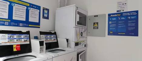 A laundry facility with multiple washing machines and dryers, a payment terminal, and informational posters on the walls.