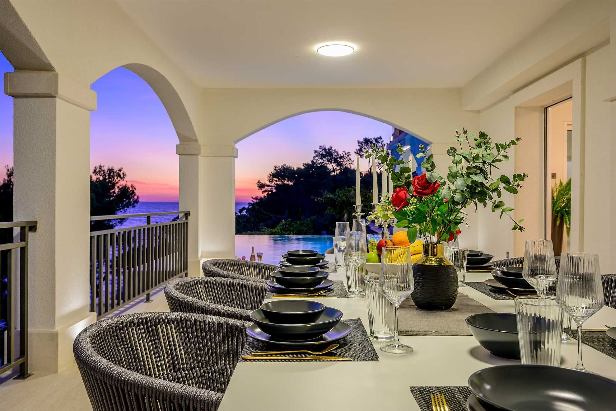 A dining area with a view of the sunset over the water, equipped with wicker chairs, black dinnerware, and a vase with flowers.
