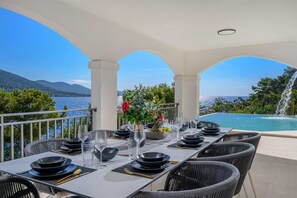 A dining area with a view of a lake and mountains, featuring a table set for four with black plates and clear glasses. - Beachfront Villa Crystal Bay with Pool (Čara)