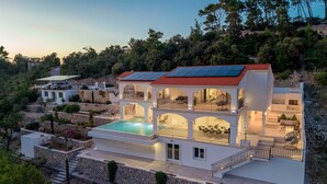 Una villa de varios pisos con piscina, mobiliario exterior y techo de tejas rojas.
