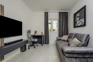 A modern living room with a flat-screen TV, a grey sofa, a desk with a chair, and a window with curtains. - Beachfront Villa Crystal Bay with Pool (Čara)